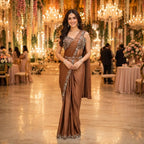 A woman stands in a grand, warmly lit event hall wearing a brown satin saree with ornate silver embroidery along the border and bodice. The background features hanging floral decorations, chandeliers, and tables with pink floral arrangements.