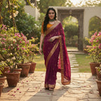 A woman wearing a deep purple saree with gold zari borders and a yellow-orange blouse, standing in a garden courtyard. The brand logo 'Stavalli Dhawan DN-03' is visible in the top right corner.