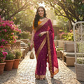 A woman stands outdoors on a stone pathway wearing a deep magenta-purple saree with gold zari embroidery and a bright orange blouse. The scene includes potted pink flowers, wicker furniture, and trees in the background, with a 'Stavvi Designer Sarees' logo in the top right corner.