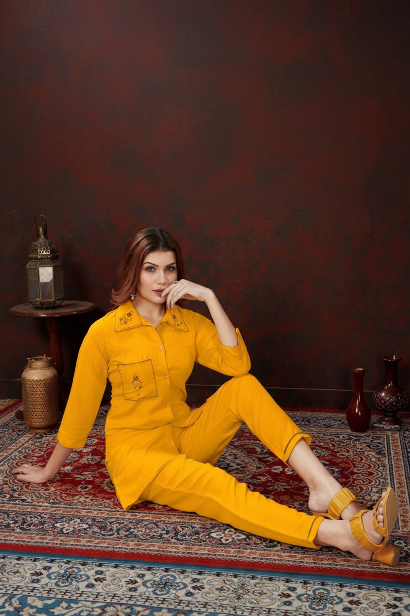 A woman sits on a patterned rug wearing a bright yellow two-piece matching set consisting of a long-sleeve button-front top and trousers, styled in an indoor setting with decorative vases and a lantern.