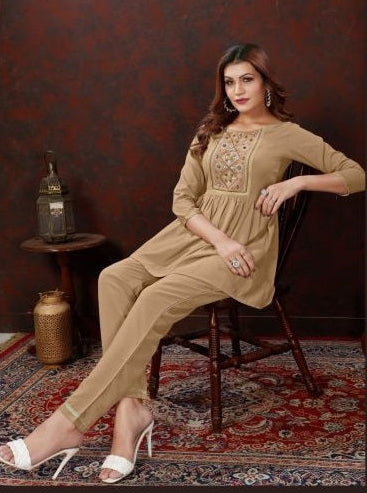 A woman models a beige three-piece kurta set consisting of a long-sleeve top with embroidered neckline, matching trousers, and white heeled sandals, seated on a wooden chair against a dark patterned wall and red rug.
