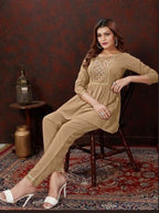 A woman models a beige three-piece kurta set consisting of a long-sleeve top with embroidered neckline, matching trousers, and white heeled sandals, seated on a wooden chair against a dark patterned wall and red rug.