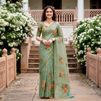 A woman stands outdoors in a courtyard wearing a sage green saree with orange and yellow floral prints. The background features stone steps, white flowering bushes, and a traditional building.