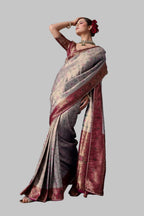 A woman models a traditional Indian saree in a studio setting with a textured gray concrete wall background. The saree has a metallic silver-gray body with a deep red border and gold zari detailing.