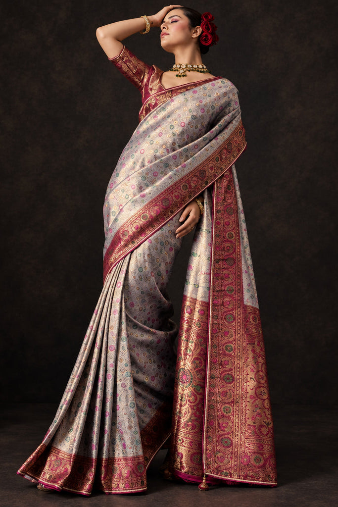 A woman models a traditional Indian saree in a studio setting with a textured gray concrete wall background. The saree has a metallic silver-gray body with a deep red border and gold zari detailing.