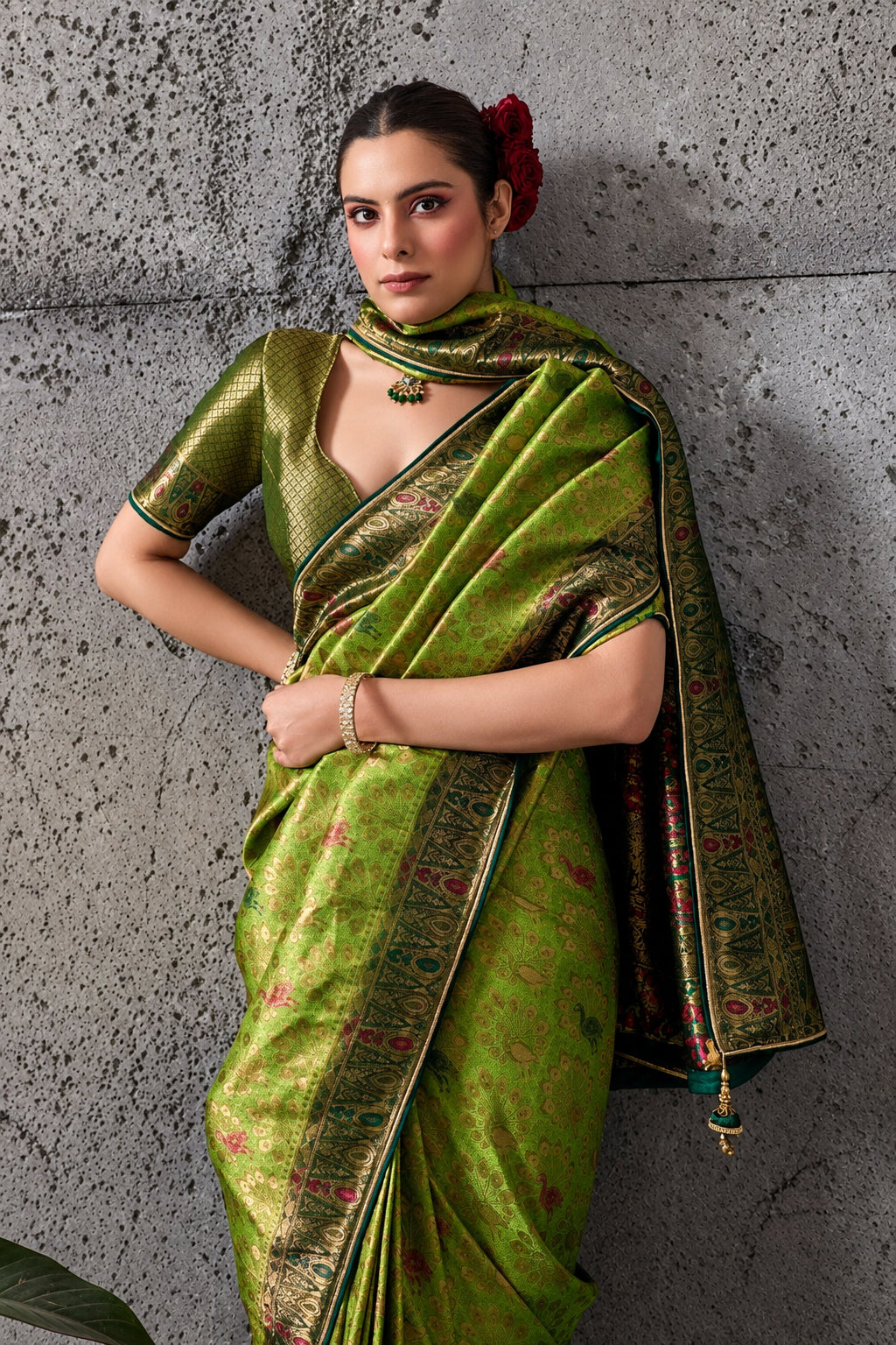 A woman models a green silk saree with gold zari embroidery and a matching blouse, standing against a textured gray concrete wall. The brand logo 'Sathvik Lifestyle' and collection name 'RAVALI 2600' are visible in the top right corner.
