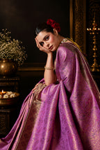 A woman wearing a purple and gold patterned sari poses against a textured gray wall. The sari has an ornate paisley-like metallic pattern and is styled with gold jewelry and a red flower in her hair.