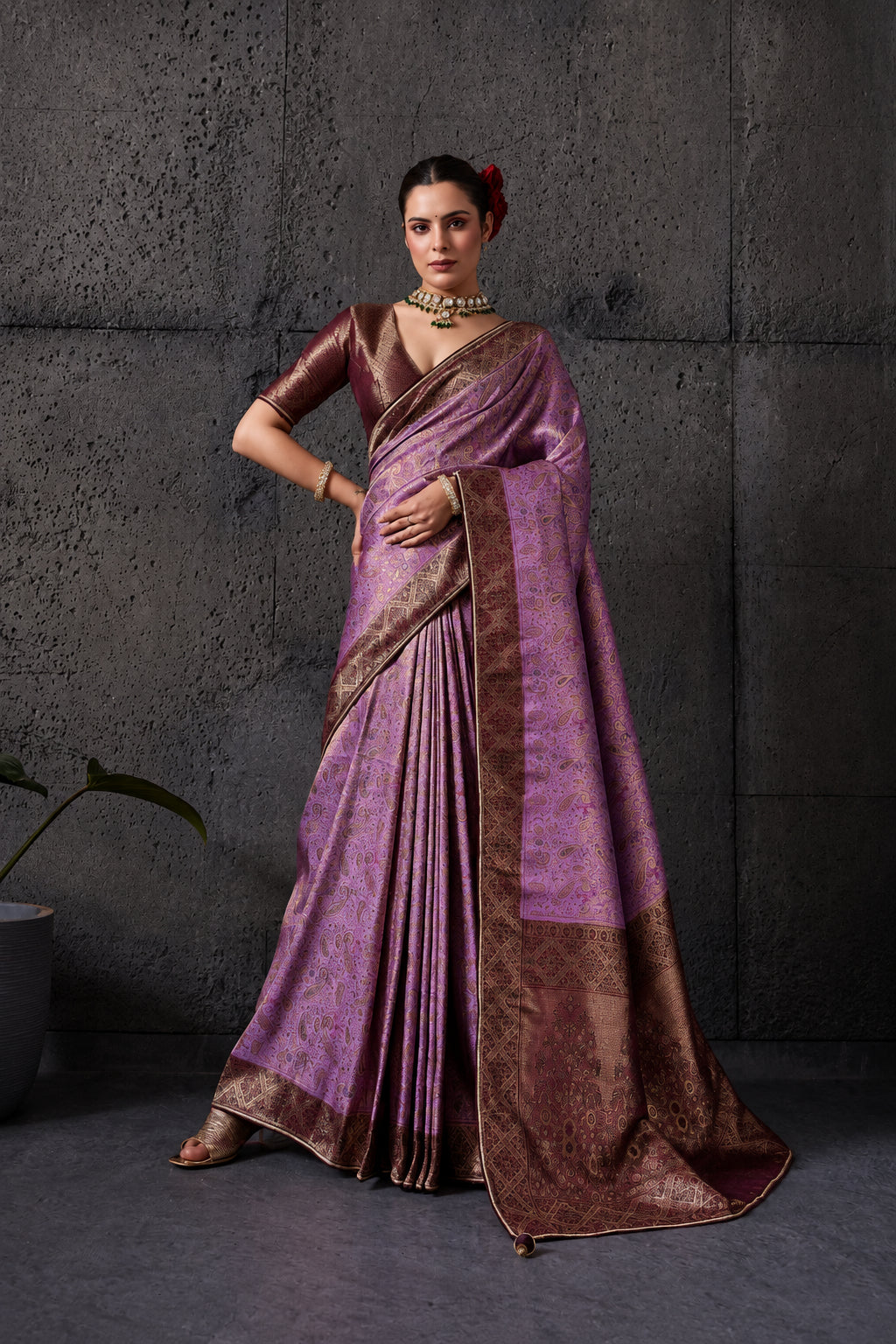 Woman wearing a pink saree with a brown border against a gray textured wall.