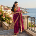 A woman wearing a deep magenta purple silk saree with gold zari borders and a gold blouse, standing on stone steps with a scenic coastal background.