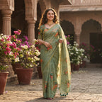 A woman stands in a courtyard wearing a sage green printed saree with floral motifs. The background features stone pillars, arches, and potted flowers.