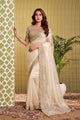 A woman models a cream-colored saree with a heavily embroidered beige blouse. The setting features a green patterned curtain on the left, a potted palm on the right, and a textured olive-green wall behind her.