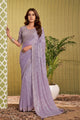 A woman models a lavender purple embroidered saree with a matching blouse, standing in front of a green patterned backdrop and a potted plant. The text 'ASK-557' is visible in the top right corner.
