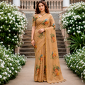 A woman stands on a stone staircase path wearing a beige printed saree with orange and green floral motifs. White flowering bushes line both sides of the walkway.