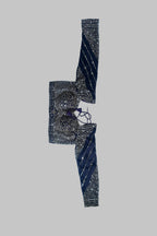 A folded traditional Indian garment, likely a sari or embroidered fabric piece, laid flat on a white sheet over a gray bedsheet. The fabric features dark navy blue borders with gold zari embroidery and motifs, and a central patterned section in black, gold, and purple.