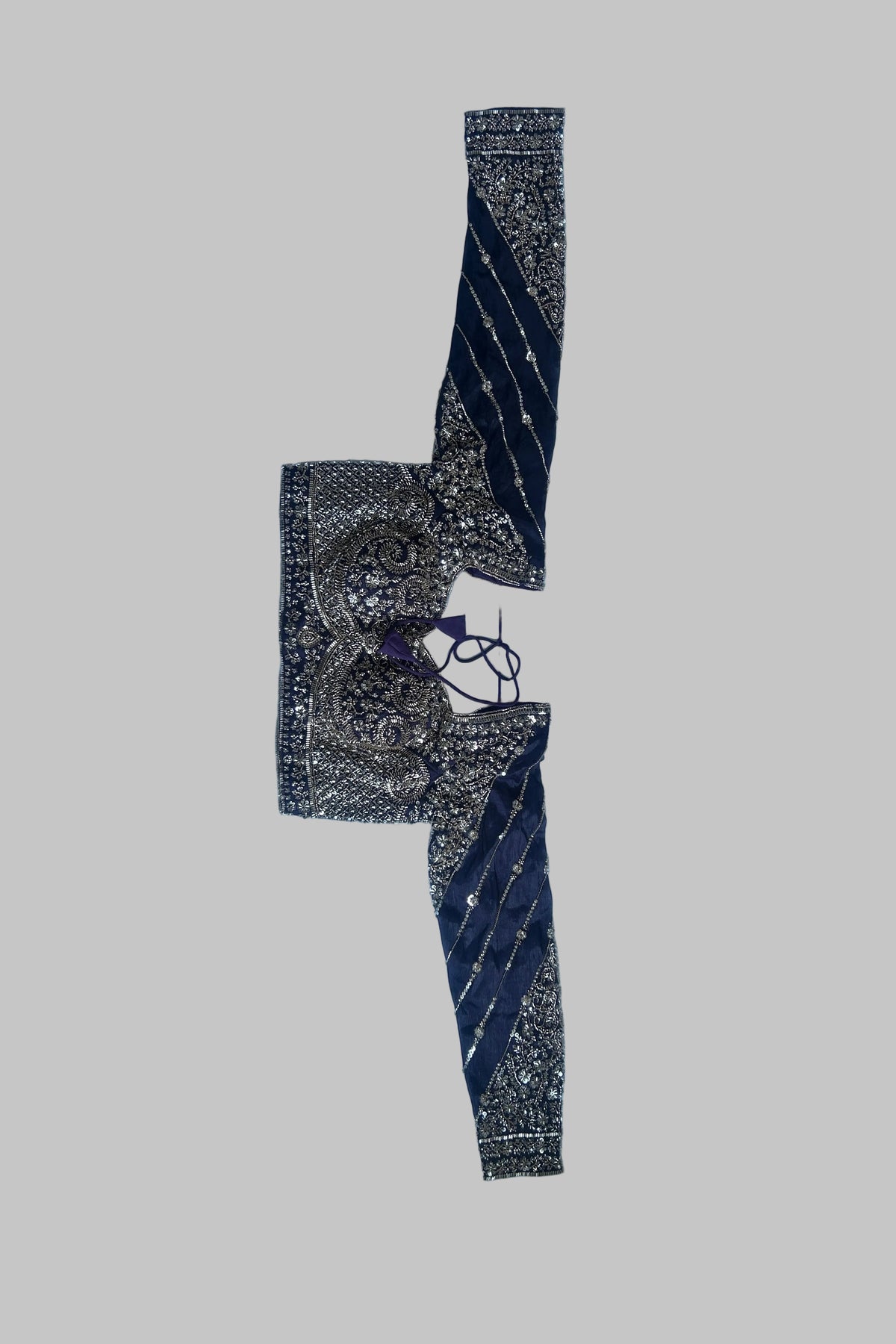 A folded traditional Indian garment, likely a sari or embroidered fabric piece, laid flat on a white sheet over a gray bedsheet. The fabric features dark navy blue borders with gold zari embroidery and motifs, and a central patterned section in black, gold, and purple.