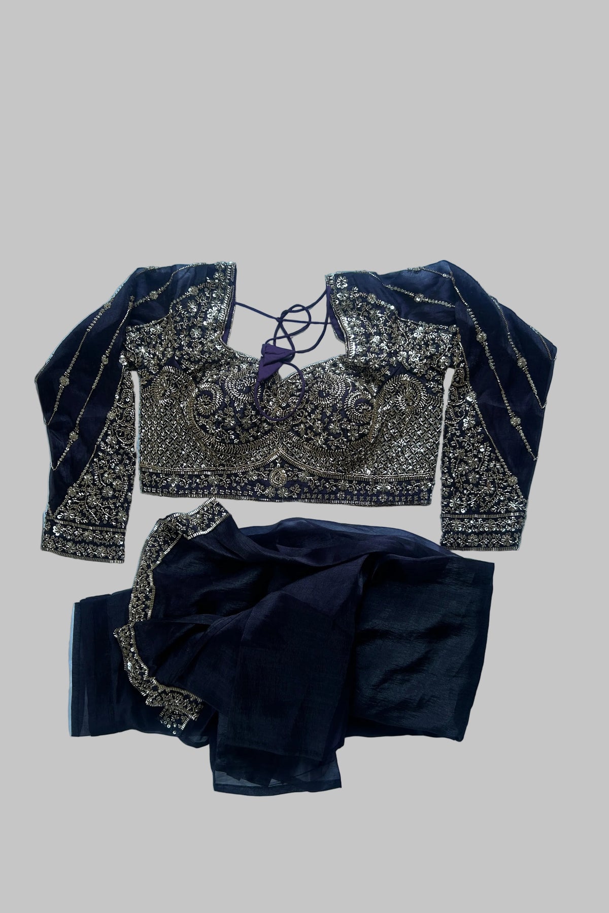 A dark navy blue traditional outfit laid flat on a white sheet, featuring an heavily embroidered blouse with gold and silver mirror work and a matching skirt.