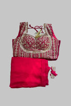 A traditional Indian outfit set featuring a deep pink/magenta blouse with intricate gold embroidery and a matching solid pink skirt, displayed on a neutral gray background.