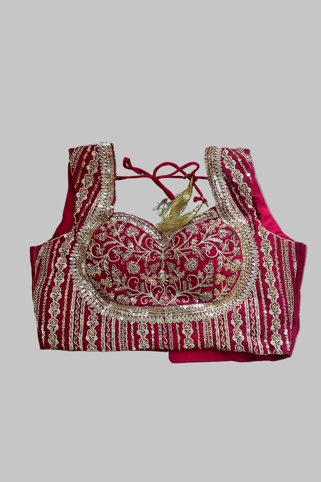 A red and gold heavily embroidered blouse top laid flat on a gray fabric background. The blouse features intricate gold sequin and thread embroidery over a deep red base, with a red tie-back neckline and a small gold tassel detail.