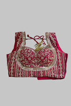 A red and gold heavily embroidered blouse top laid flat on a gray fabric background. The blouse features intricate gold sequin and thread embroidery over a deep red base, with a red tie-back neckline and a small gold tassel detail.