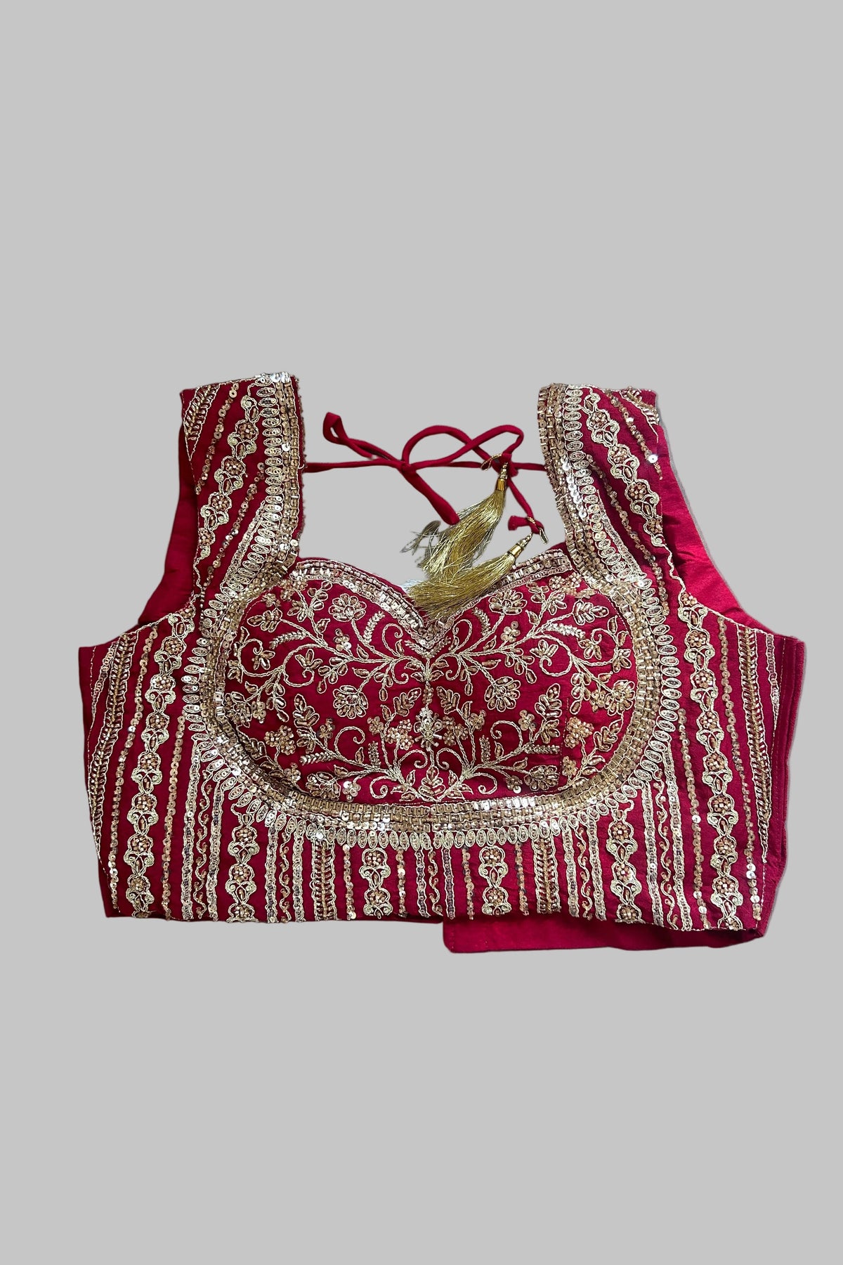 A red and gold heavily embroidered blouse top laid flat on a gray fabric background. The blouse features intricate gold sequin and thread embroidery over a deep red base, with a red tie-back neckline and a small gold tassel detail.