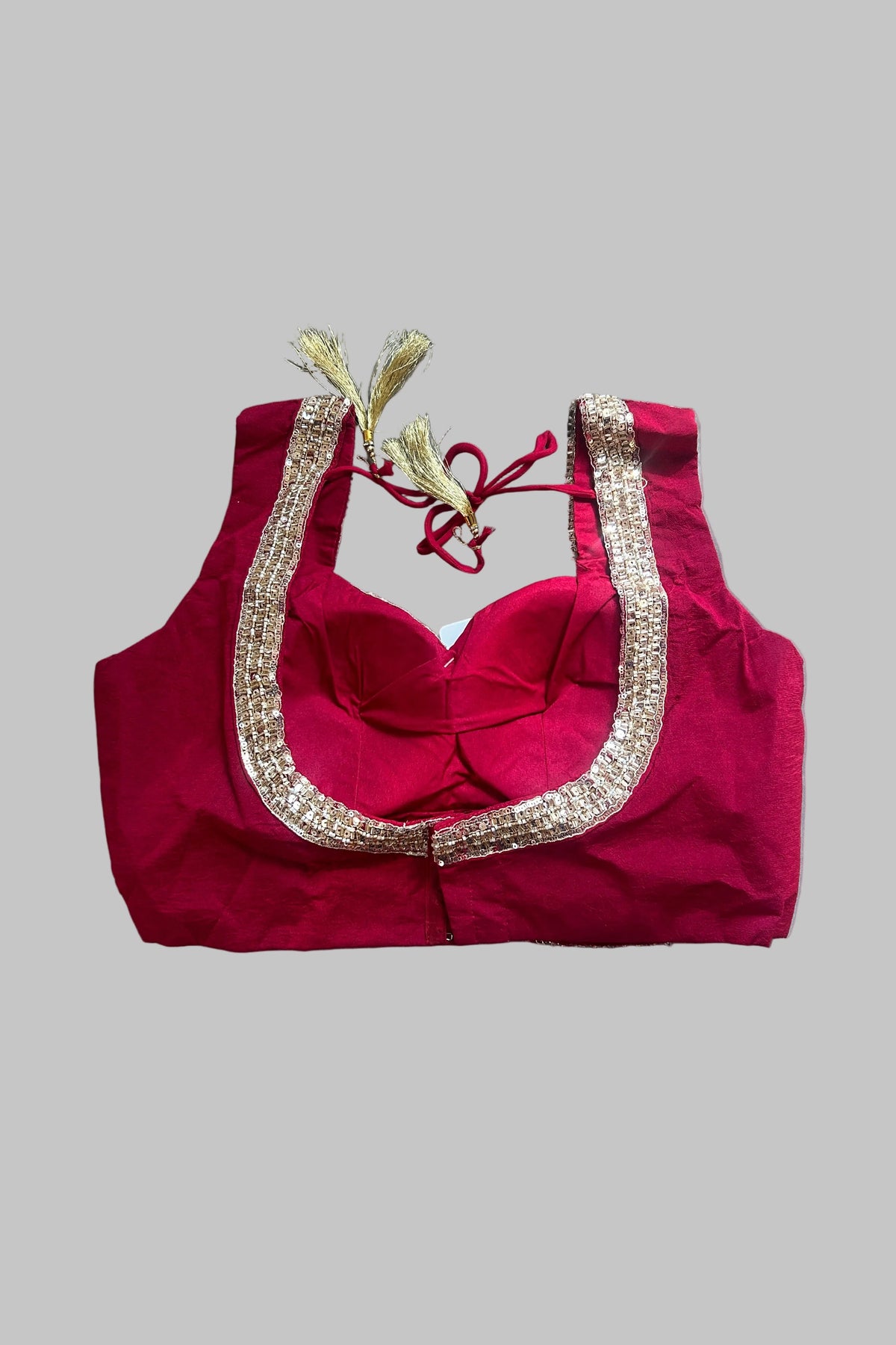 A bright pinkish-red sleeveless blouse top with a sweetheart neckline, decorated with a wide gold sequined border and red tie strings with gold tassels, laid flat on a gray fabric background.