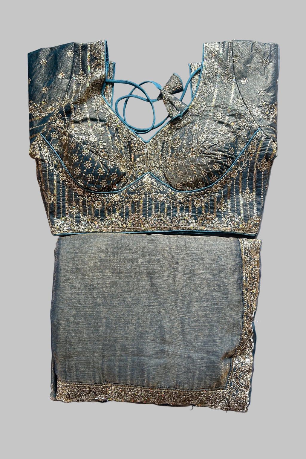 A traditional Indian blouse and skirt set laid flat on a gray fabric background. The blouse is made of teal blue fabric with intricate gold zari embroidery and lace-up neck detailing.