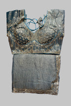 A traditional Indian blouse and skirt set laid flat on a gray fabric background. The blouse is made of teal blue fabric with intricate gold zari embroidery and lace-up neck detailing.