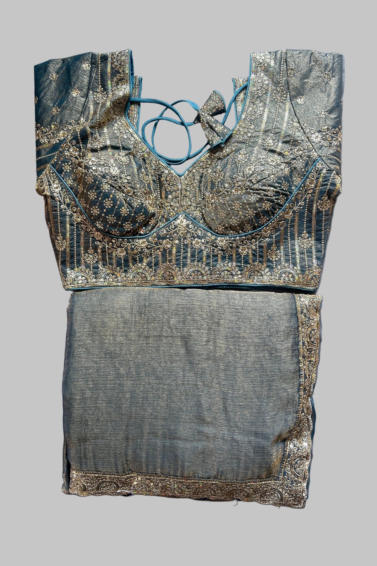 A traditional Indian blouse and skirt set laid flat on a gray fabric background. The blouse is made of teal blue fabric with intricate gold zari embroidery and lace-up neck detailing.