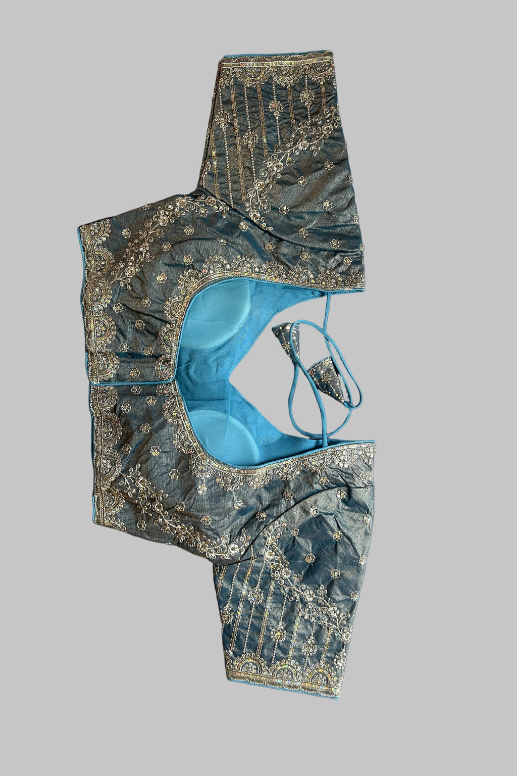 A blue and gold embroidered blouse piece laid flat on a gray fabric background, showing a light blue inner lining and decorative gold metallic embroidery.