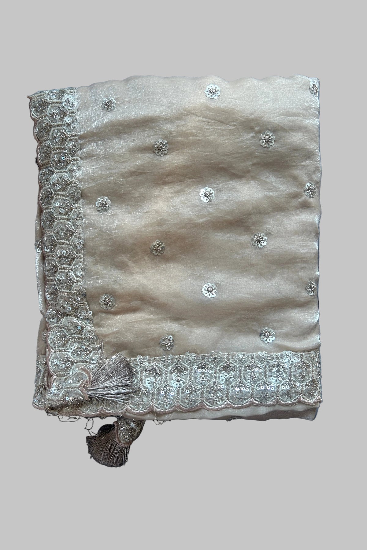 A folded beige fabric item, likely a dupatta or scarf, decorated with silver sequins and a detailed silver embroidered border, laid flat on a gray fabric background.