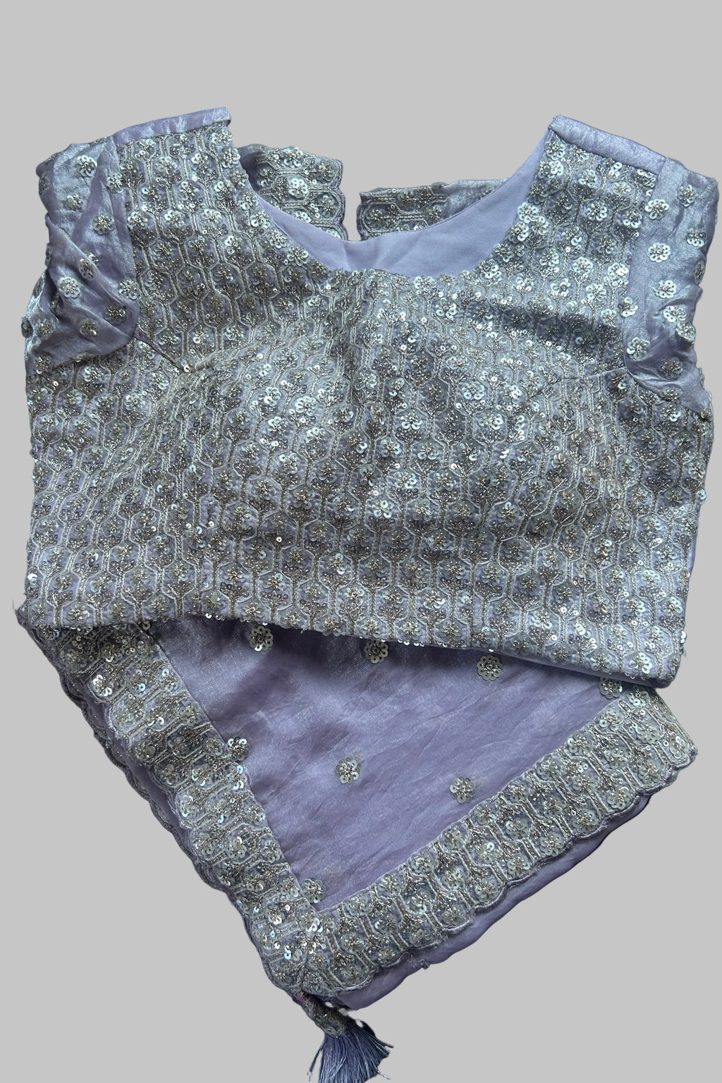 A lavender purple and silver embellished traditional Indian outfit laid flat on a dark gray fabric background. The top features intricate silver embroidery and beadwork over a lavender base, paired with a matching lavender bottom piece decorated with silver border embroidery.