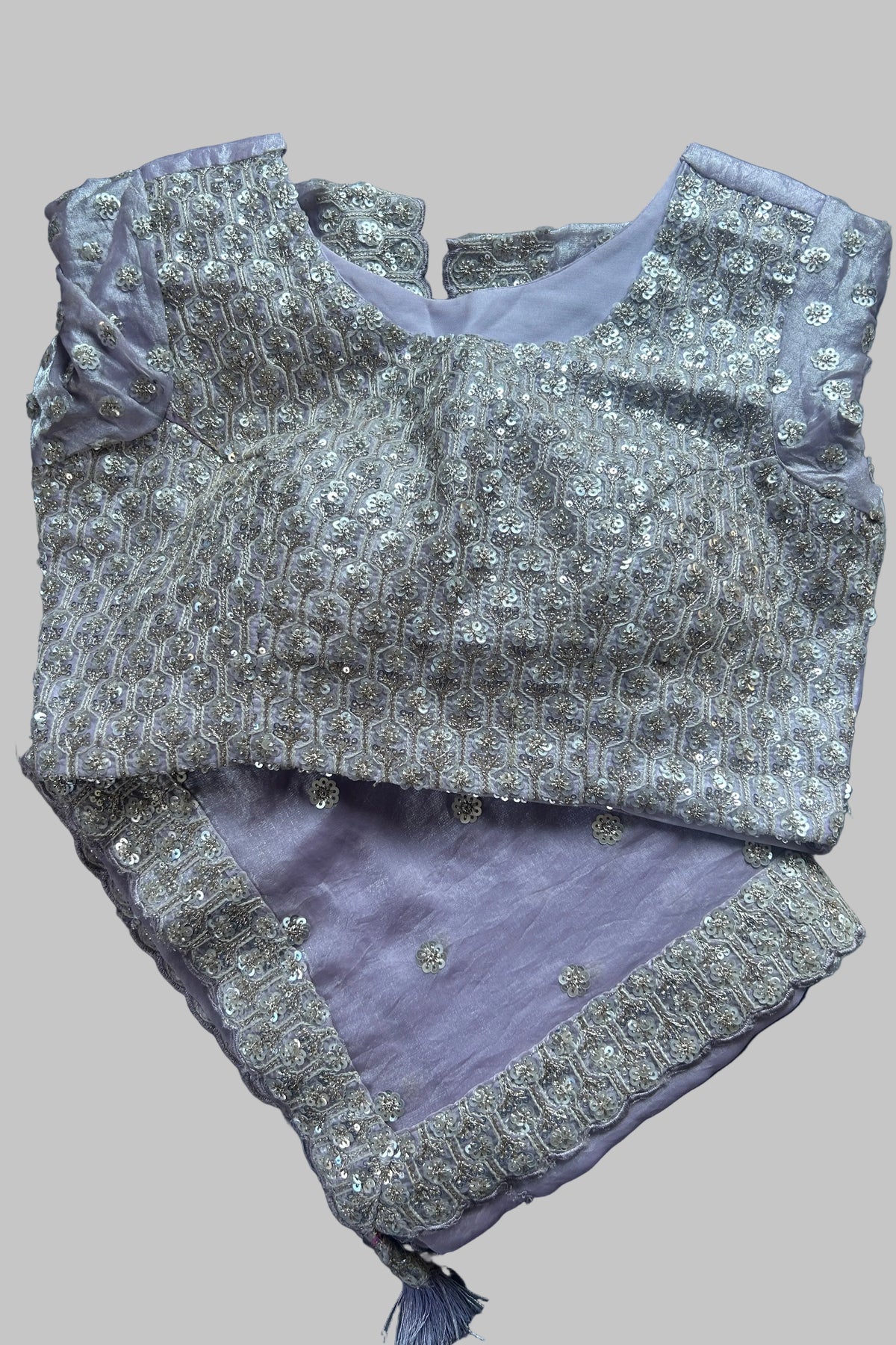 A lavender purple and silver embellished traditional Indian outfit laid flat on a dark gray fabric background. The top features intricate silver embroidery and beadwork over a lavender base, paired with a matching lavender bottom piece decorated with silver border embroidery.
