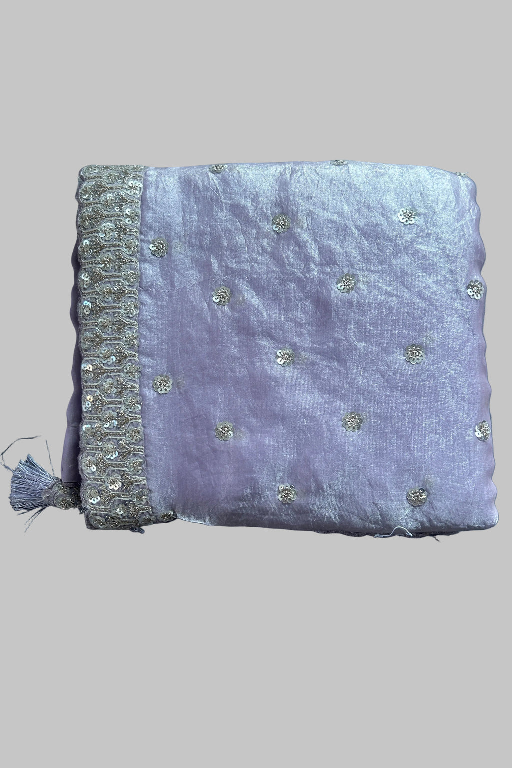 A folded lavender purple fabric item, likely a dupatta or scarf, featuring delicate silver sequin embroidery and a decorative silver border with a tassel, laid flat on a gray fabric background.