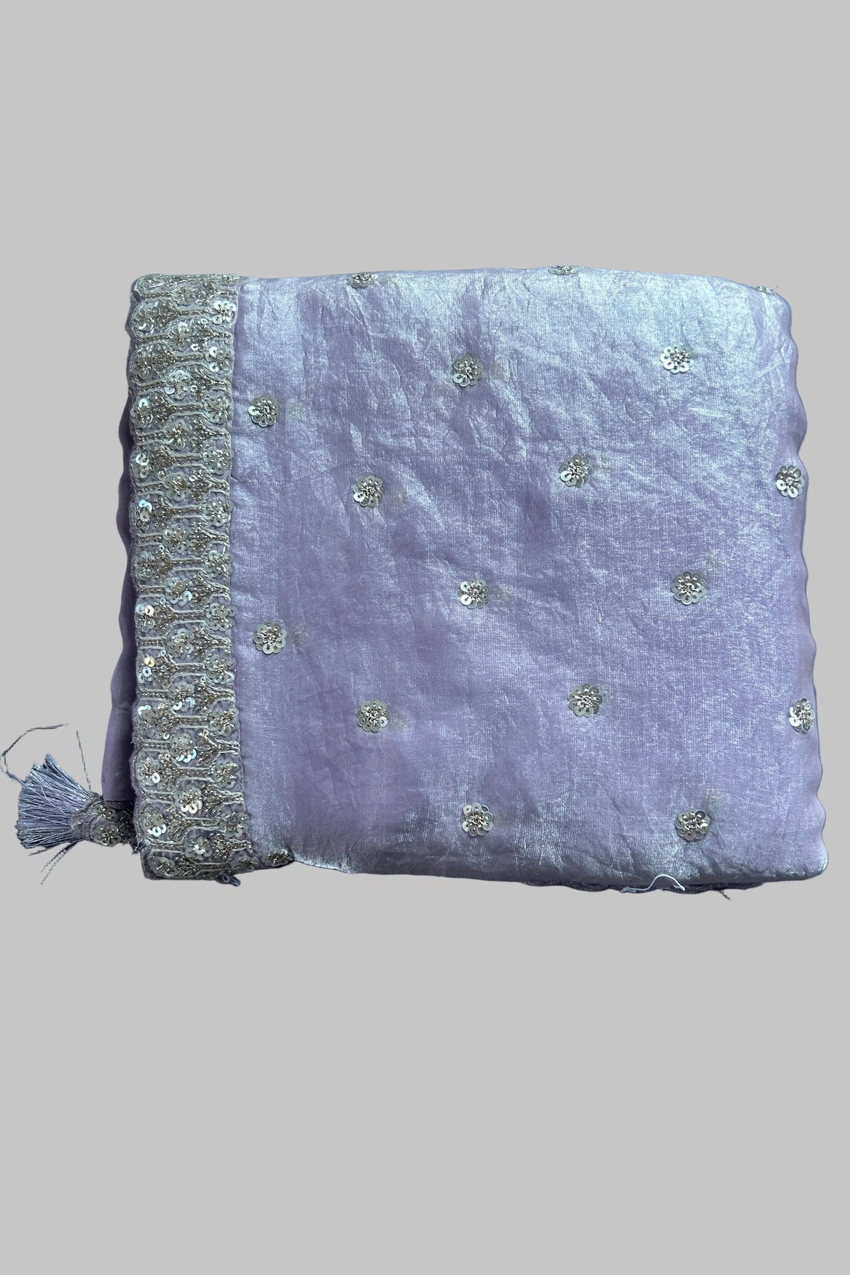 A folded lavender purple fabric item, likely a dupatta or scarf, featuring delicate silver sequin embroidery and a decorative silver border with a tassel, laid flat on a gray fabric background.