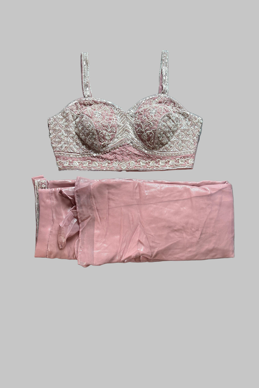 A two-piece lingerie set laid flat on a gray bedsheet, featuring a light pink and white floral lace bralette with matching pink satin pants. A person's feet and sandals are visible at the edges of the frame.