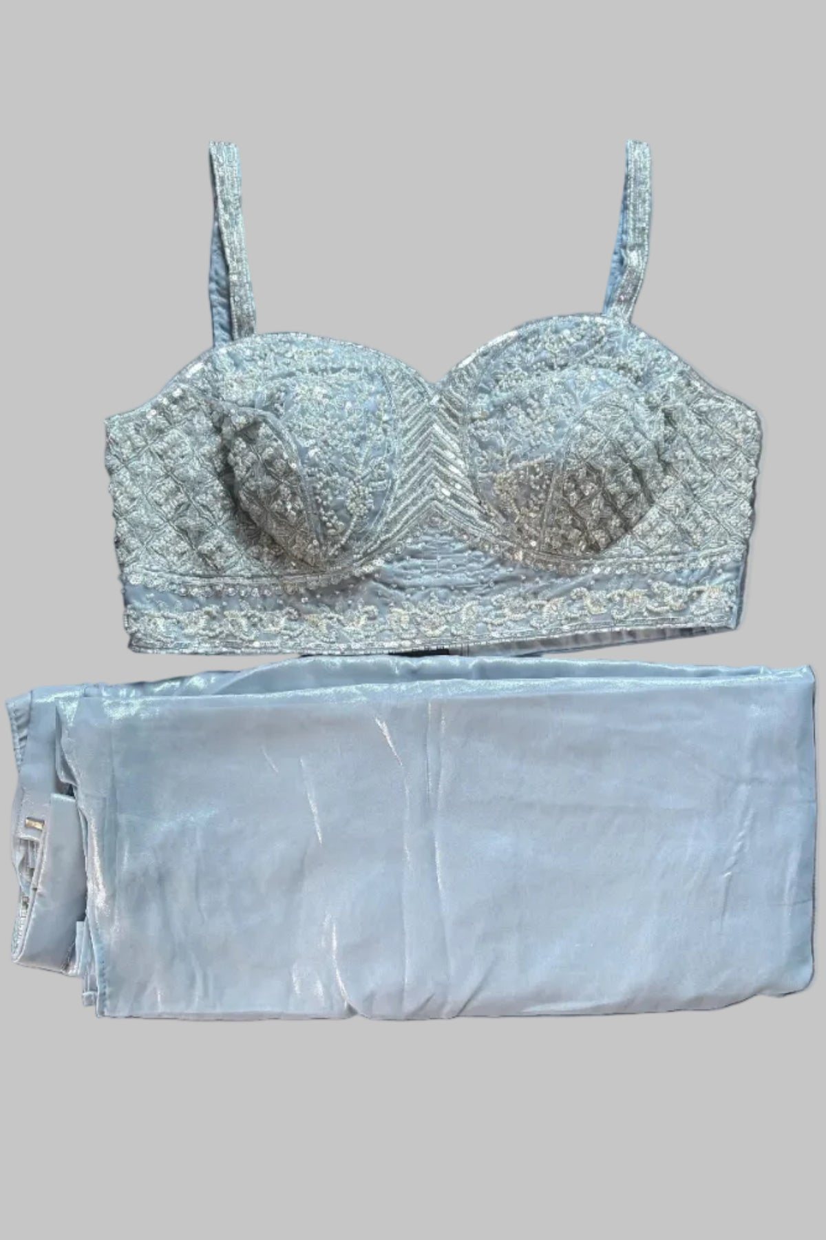 A light blue lace and embroidered bralette laid flat on a gray fabric surface, with matching folded light blue pants or a skirt placed underneath it.