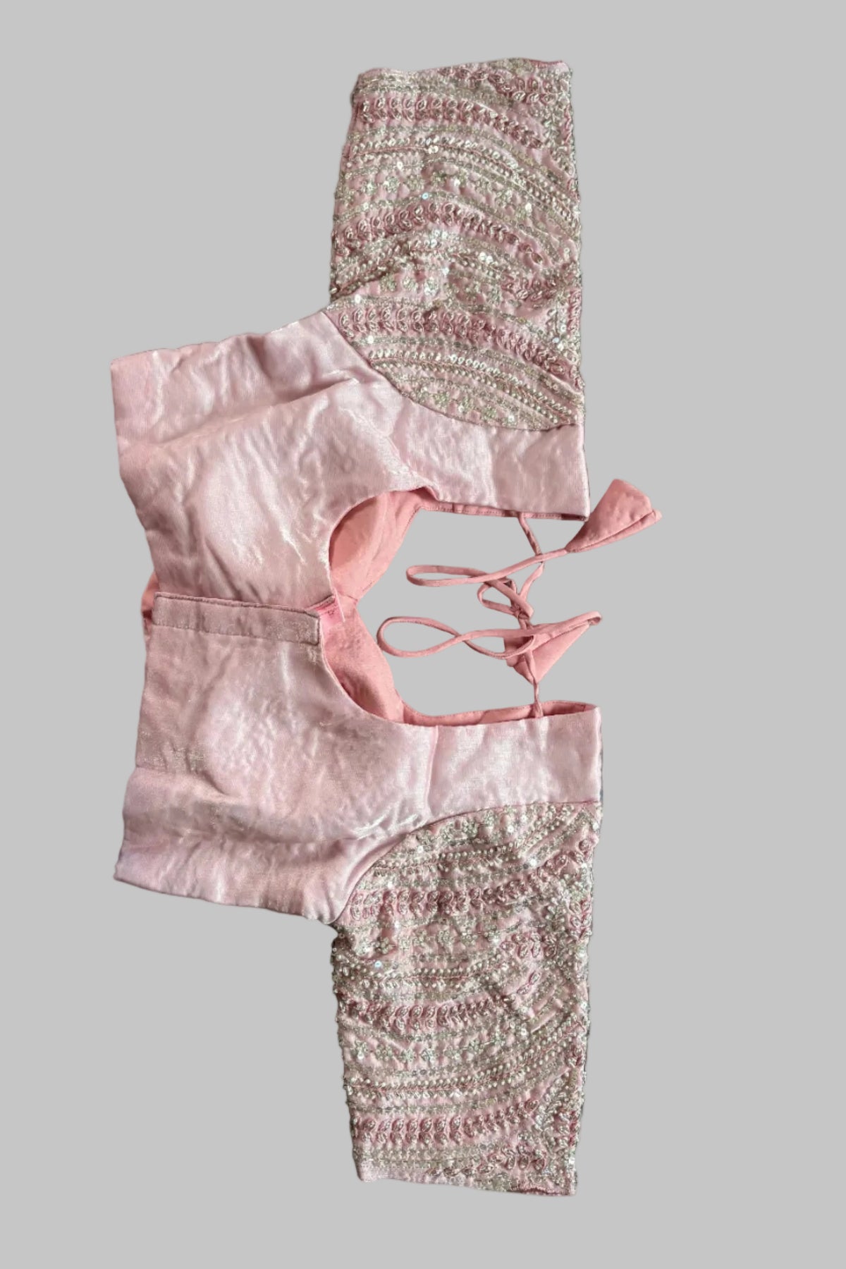 A light pink blouse top with intricate silver and gold embroidery on the sleeves and a lace-up back detail, laid flat on a gray fabric surface.