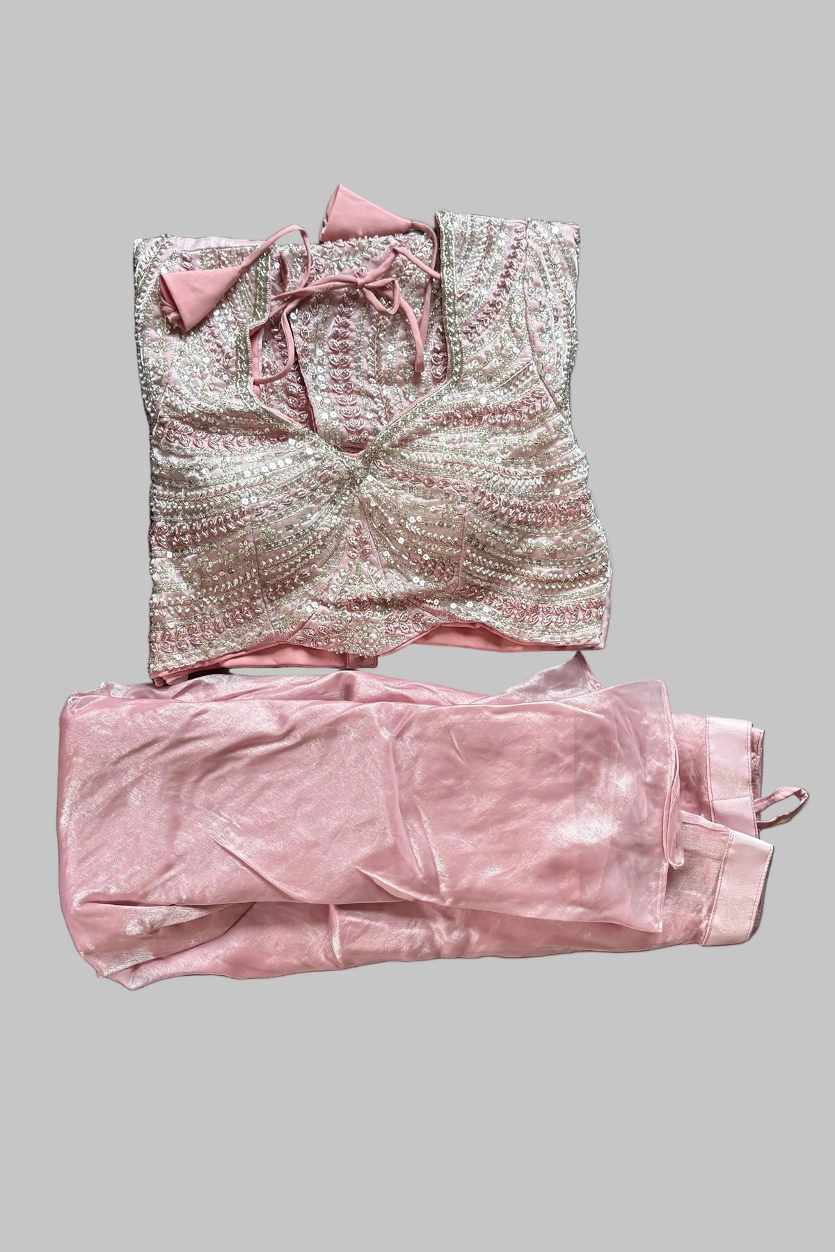 A folded two-piece clothing set laid flat on a gray bedsheet. The set includes a light pink and white patterned top with a bow at the neckline and matching light pink pants.