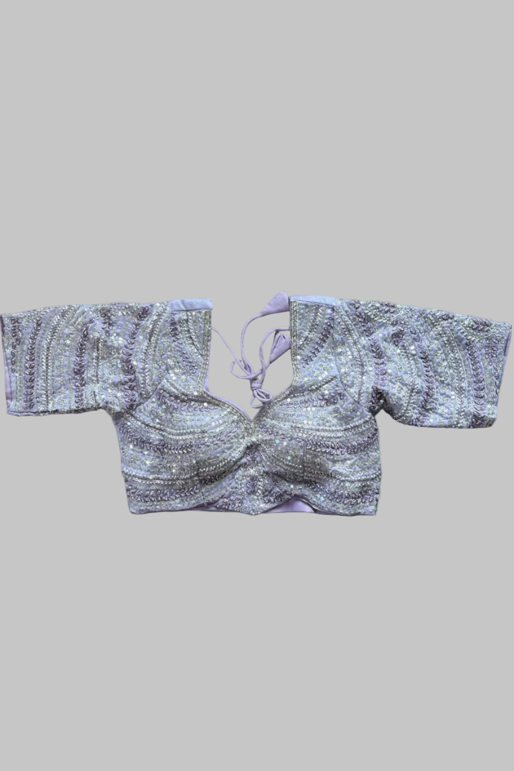 A lavender and silver embellished crop blouse with wide sleeves and a tie neckline, laid flat on a gray bed with feet visible at the bottom corners.