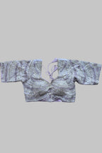 A lavender and silver embellished crop blouse with wide sleeves and a tie neckline, laid flat on a gray bed with feet visible at the bottom corners.