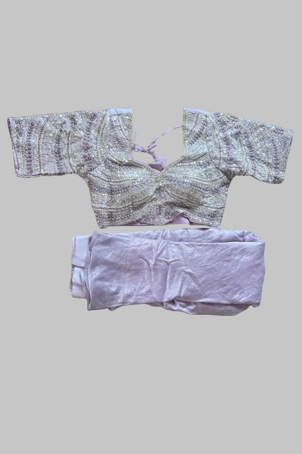 A folded traditional Indian outfit laid flat on a wrinkled gray sheet, featuring a heavily embroidered light pink and gold blouse with a tie-neck and matching light pink pants.