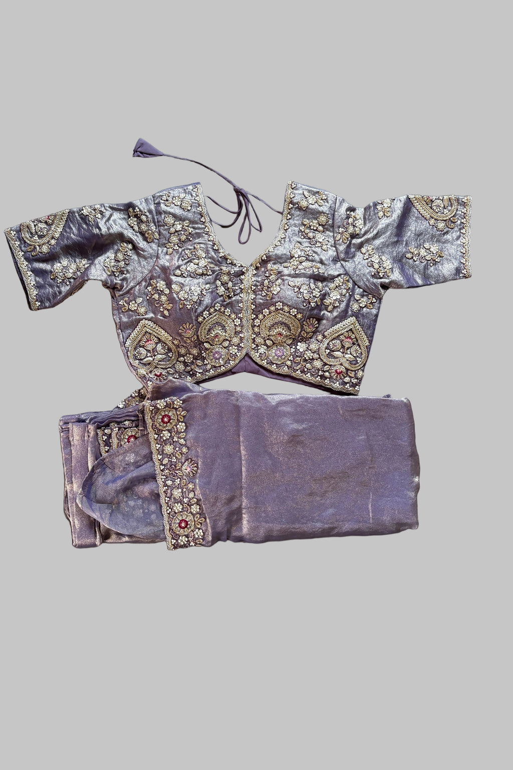 A lavender purple traditional Indian ethnic wear set, including a heavily embroidered kurta and matching trousers, laid flat on a gray bedsheet next to a folded matching fabric piece.