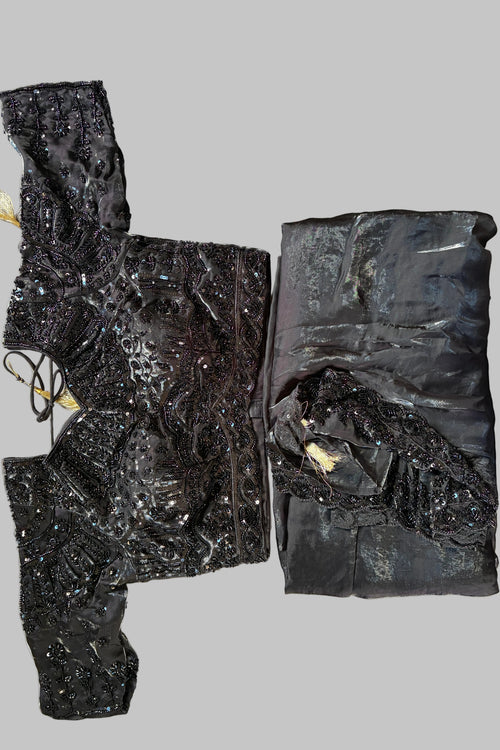 A black embellished traditional garment laid flat on a white satin fabric background, showing a folded matching fabric piece alongside the main decorated top and sleeves with sequins and beadwork.