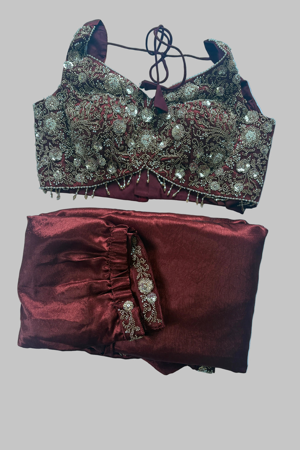 A traditional Indian ethnic wear set consisting of a heavily embroidered maroon and gold blouse with a matching tie-neck cord and folded maroon skirt with a coordinating embroidered border, laid flat on a white sheet.