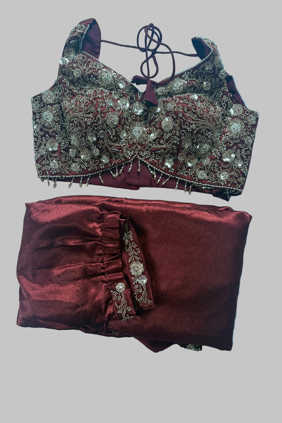 A traditional Indian ethnic wear set consisting of a heavily embroidered maroon and gold blouse with a matching tie-neck cord and folded maroon skirt with a coordinating embroidered border, laid flat on a white sheet.