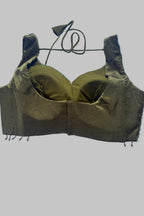 An olive green fabric bralette or underwire bra laid flat on a light blue wrinkled sheet. The garment has a textured shimmering fabric, a center seam, and a thin matching cord tie at the top.