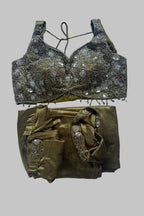 A two-piece traditional Indian outfit laid flat on a light blue fabric background. The set includes a metallic gold and bronze embellished blouse with silver mirror work and a matching pleated skirt with embroidered details.