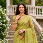 A woman wearing a light olive green saree with orange floral patterns and gold jewelry, standing on stone stairs in front of a white railing with white flowers on either side.