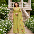 A woman stands on a stone pathway in front of a garden pavilion, wearing a light olive green saree with orange and green floral prints. The background features white flowers, green foliage, and a white stone railing.