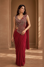A woman modeling a deep maroon red saree with an intricately embroidered sleeveless bodice in silver and red tones, set against a warm brown studio backdrop.